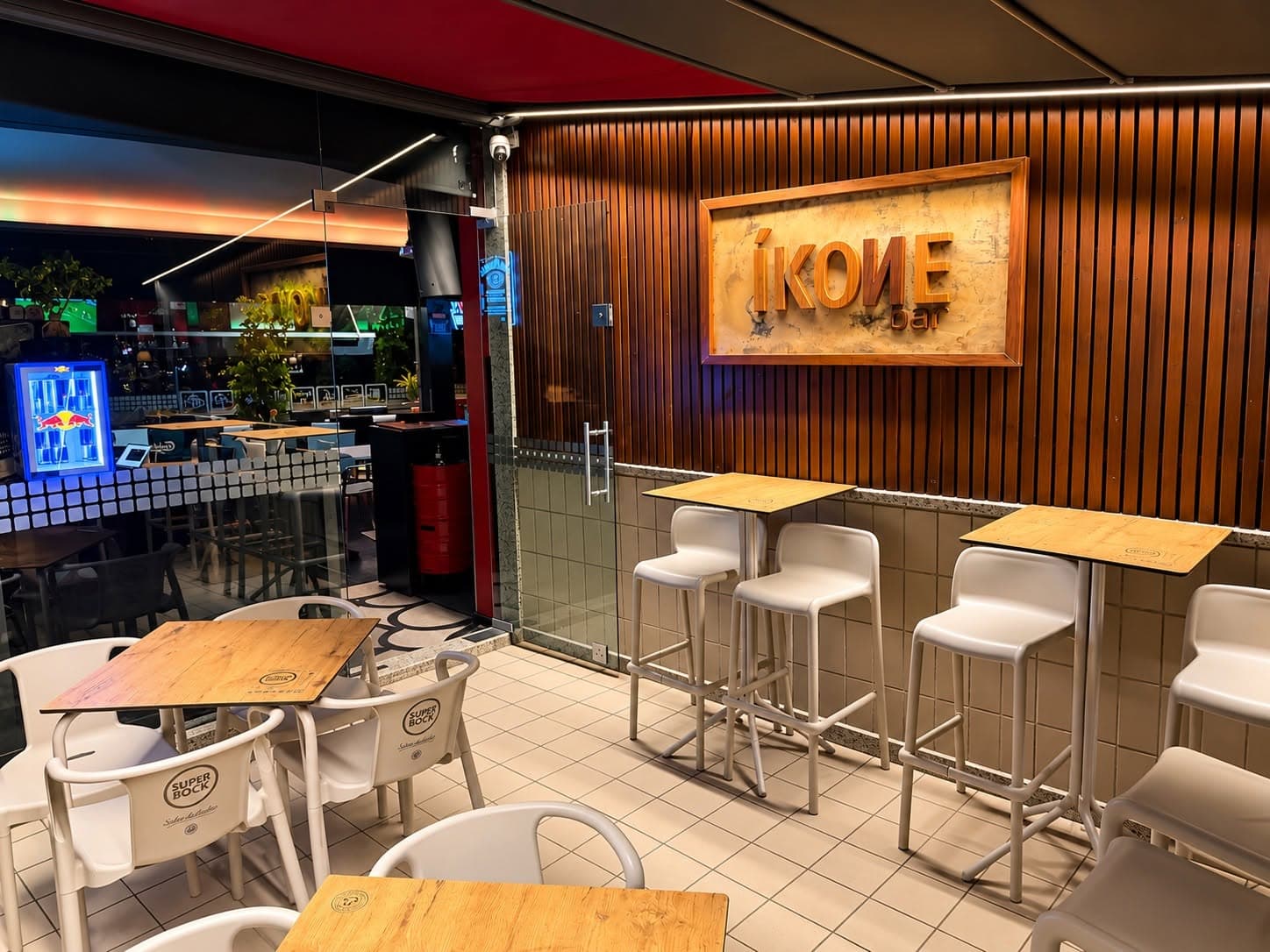Esplanada coberta do IKONE bar com mesas altas e letreiro em madeira
