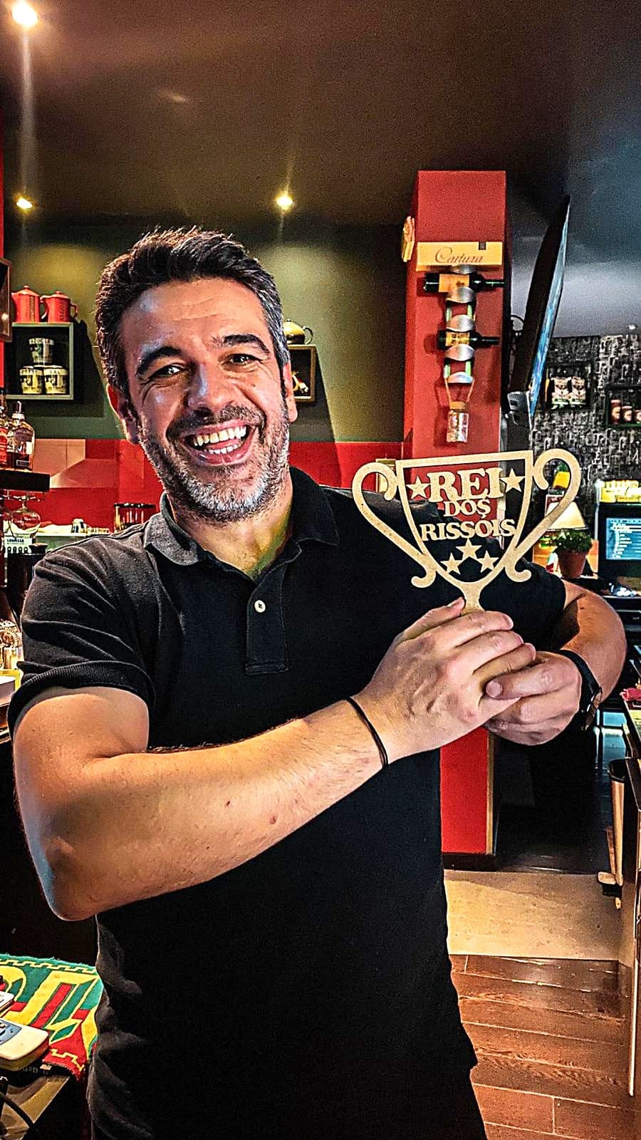 Homem sorridente segura um troféu «Rei dos Rissóis» atrás do balcão