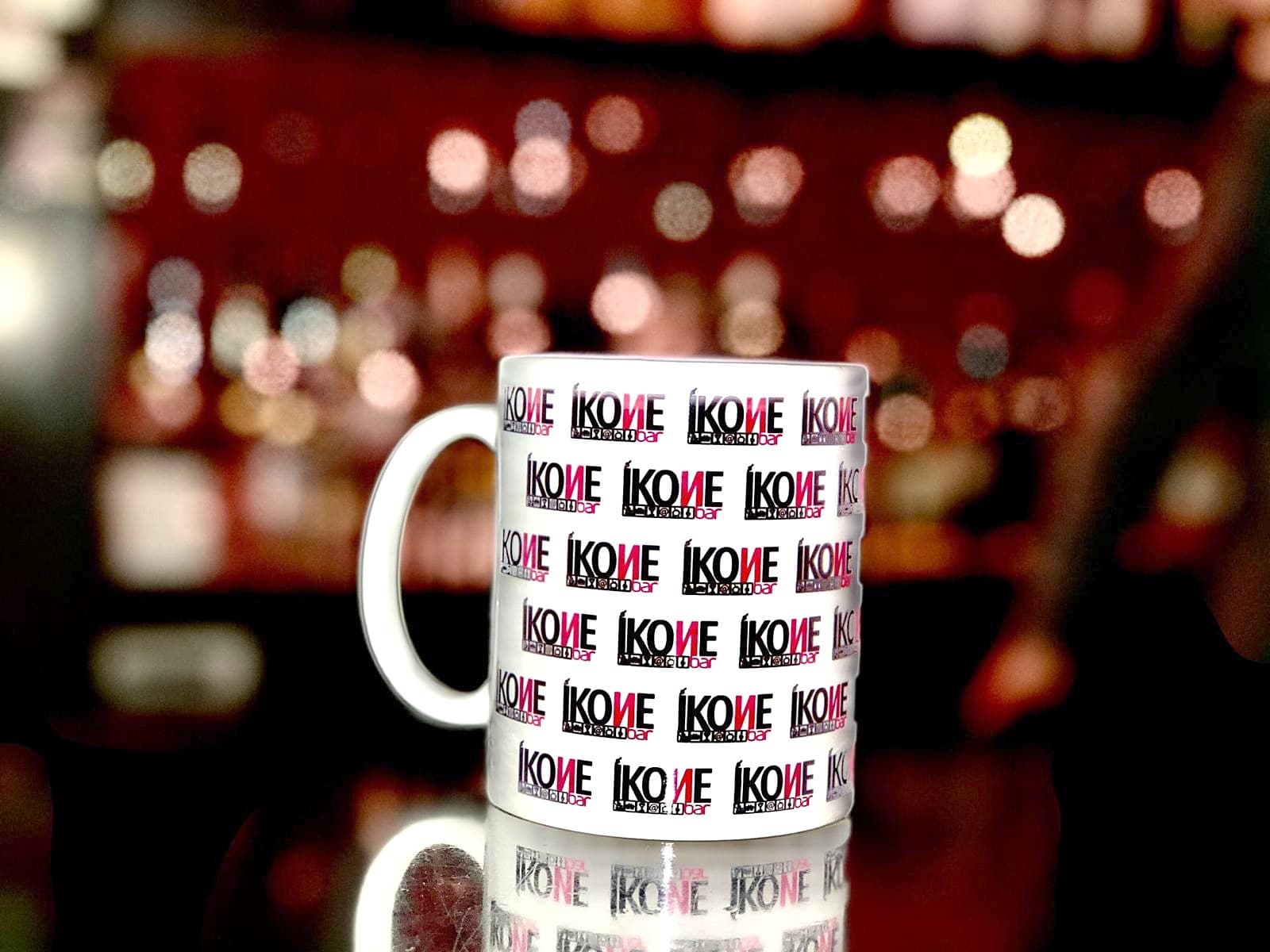 Caneca branca com o logótipo IKONE repetido, com as luzes do bar desfocadas atrás
