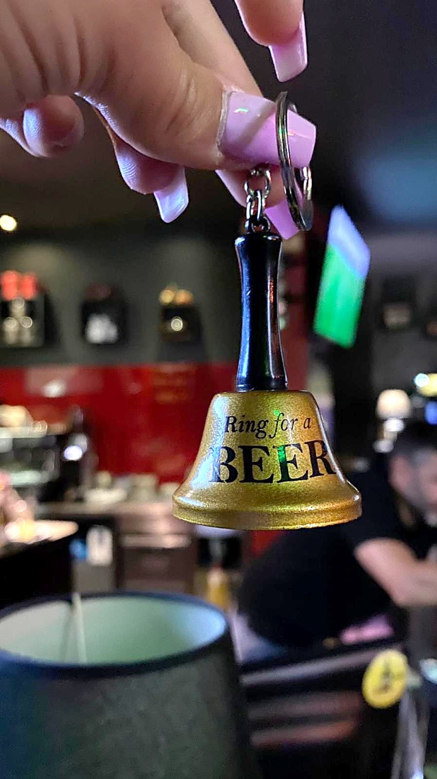 Mão a tocar um sino dourado «Ring for a beer», com a sala do bar desfocada