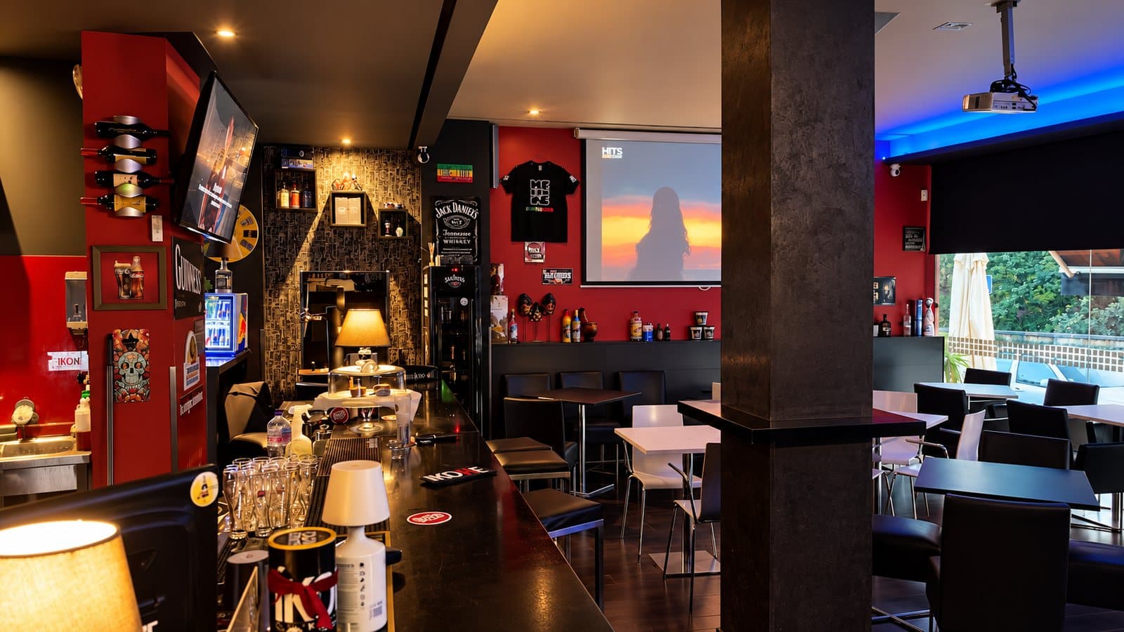 Sala do IKONE bar com balcão comprido, coluna escura, mesas e projetor ligado sobre parede vermelha