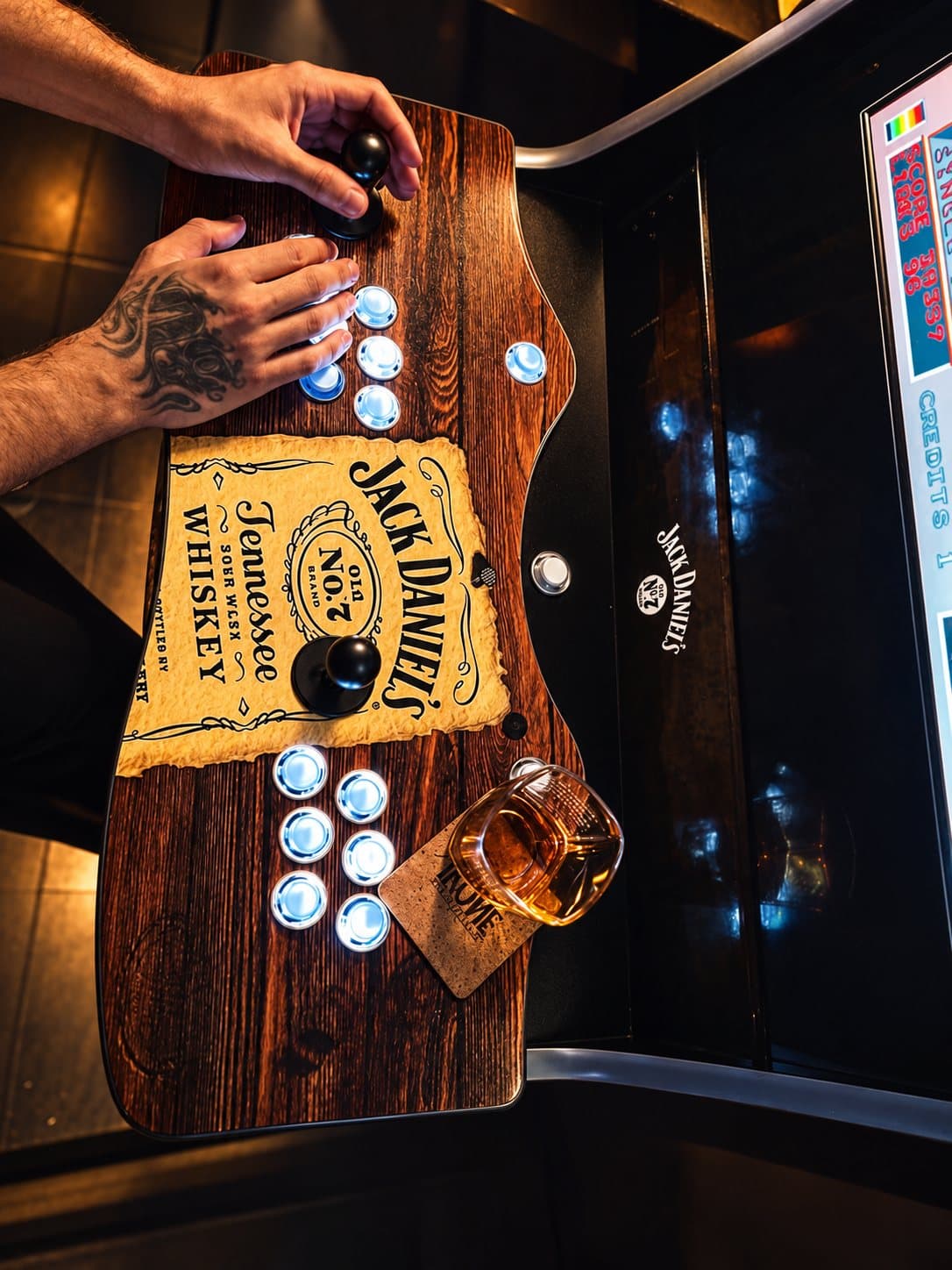 Mãos nos comandos de uma máquina arcade Jack Daniel's, com um copo de whisky ao lado